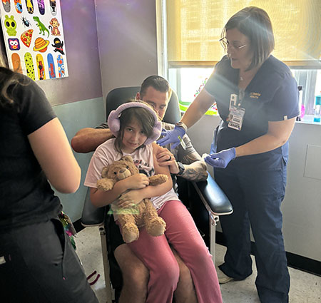 A young girl wearing fuzzy pink headphones sits in a medical chair on her father's lap while two health care providers prep her for a vaccine.