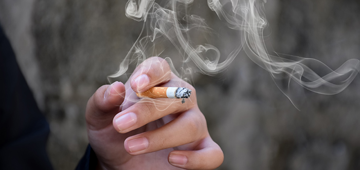 A hand holds a lit cigarette with smoke coming from it. 