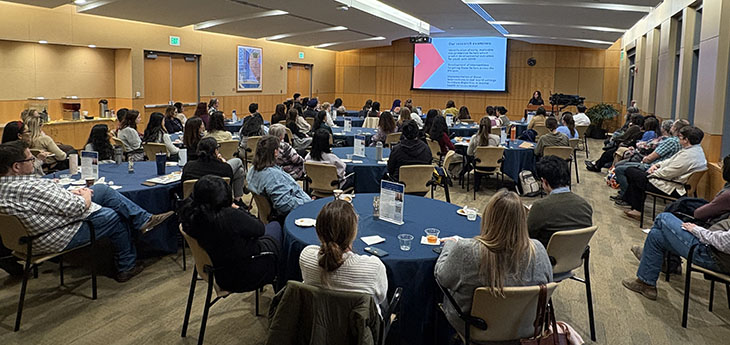 A large group of people seated in chairs at round tables listen to a speaker at the MIND Institute auditorium. 