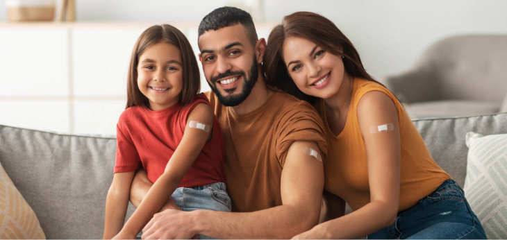 A smiling family sits on a couch with their sleeves rolled up showing an adhesive bandage on their shoulder.