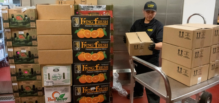 Person moves boxes of food supplies from a delivery cart inside a storage area lined with stacked produce and shipping cartons.