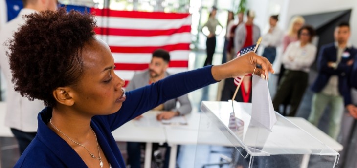 Person casts a ballot into a clear voting box with an American flag in the background.
