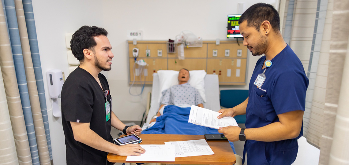Two male nursing students review patient records with a medical mannequin lying in a hospital bed behind them.