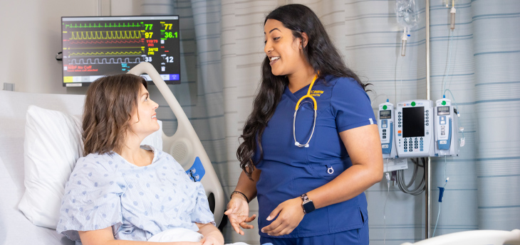 Nurse in blue scrubs smiles while speaking with a patient in a hospital bed with monitors in the background.