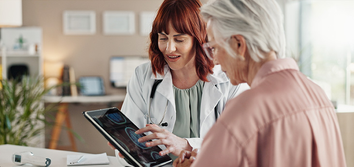 A healthcare provider in a white coat shows medical images on a tablet to an older woman during a consultation.