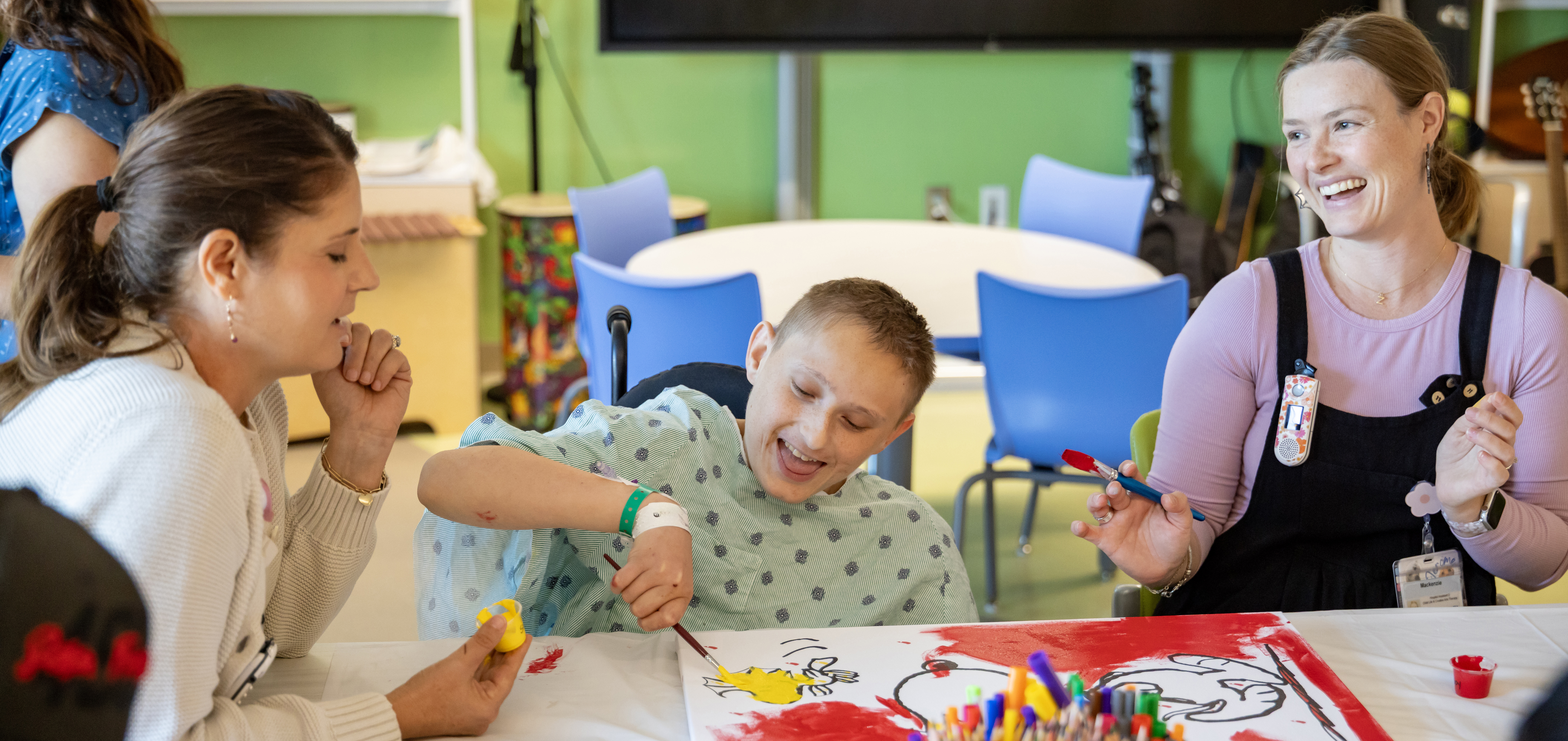 Two adults sit by child in hospital gown painting a Charlie Brown mural. 