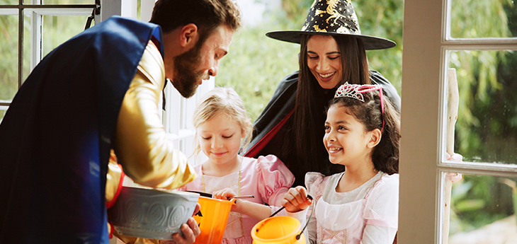 Two children and woman in witch hat stand in doorway while man reaches into bowl.