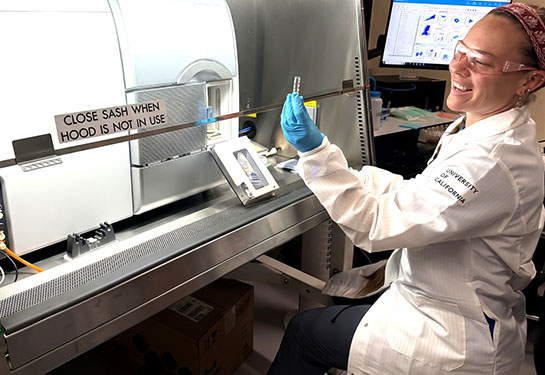 A researcher in a lab coat smiles as she holds up a test tube. 