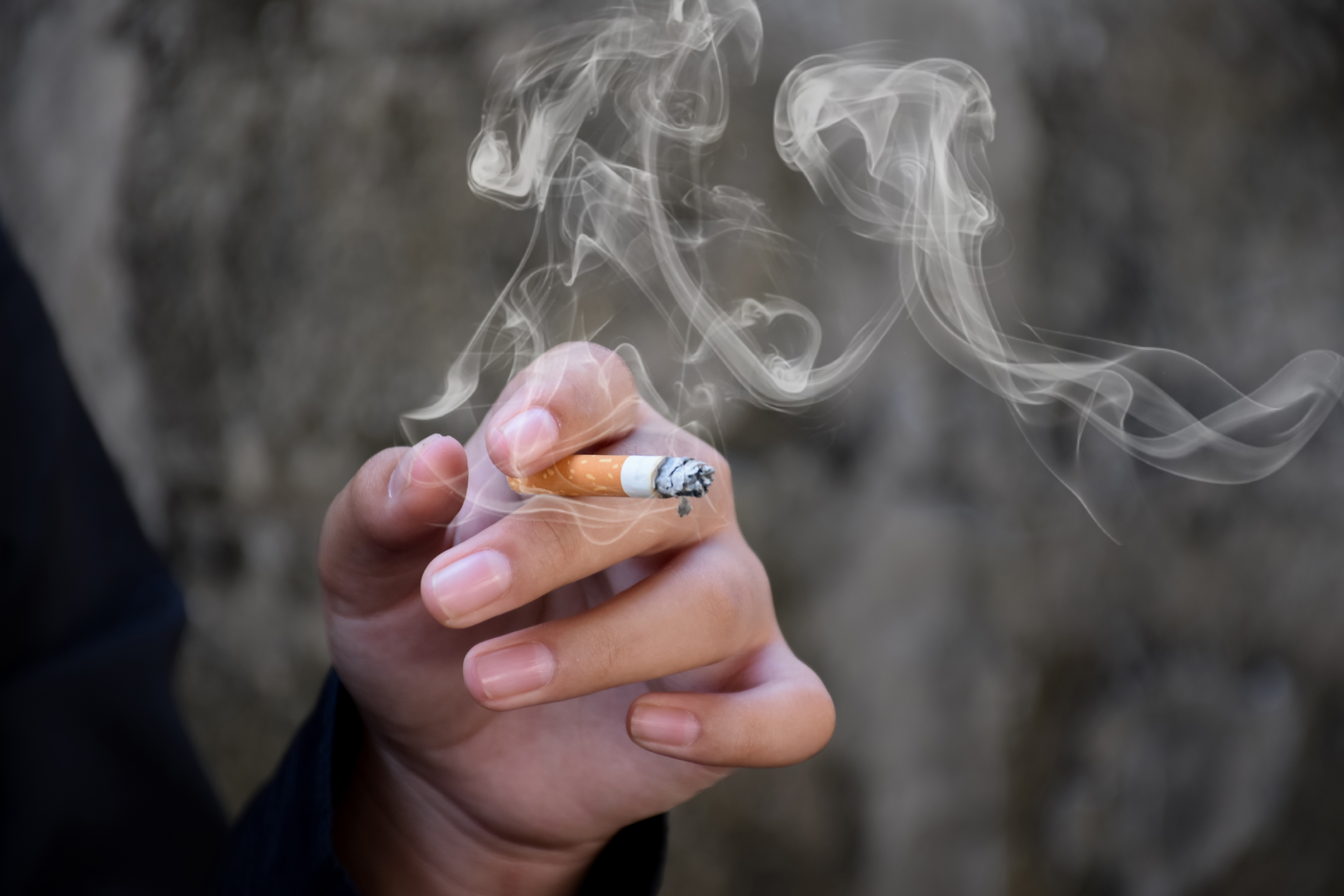 A hand holds a lit cigarette with smoke coming from it. 
