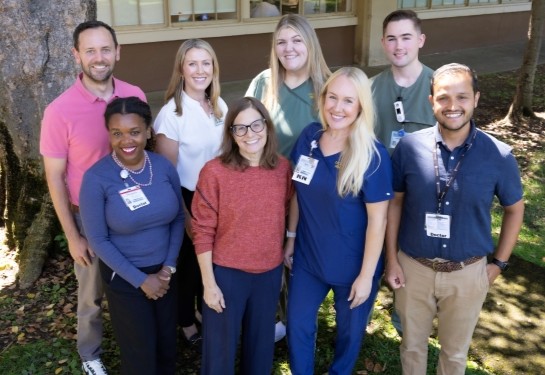 Eight smiling adults are gathered outdoors in front of a tree. 
