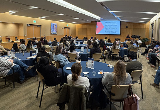 A large group of people seated in chairs at round tables listen to a speaker at the MIND Institute auditorium.
