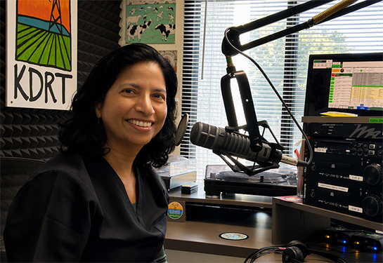 Ulfat Shaikh smiles sitting in front of a microphone in a studio. 