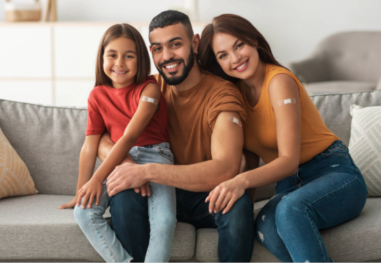A smiling family sits on a couch with their sleeves rolled up showing an adhesive bandage on their shoulder.
