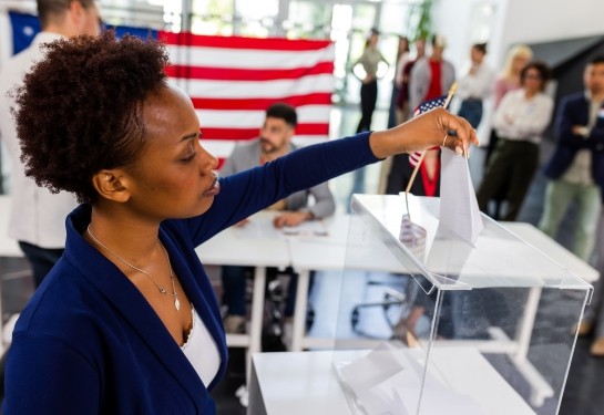 Person casts a ballot into a clear voting box with an American flag in the background.