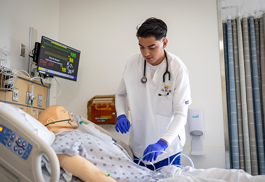 Male nursing student wearing white scrubs and blue gloves practices patient care on a mannequin in simulation lab.