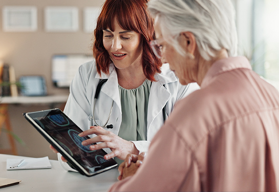 A healthcare provider in a white coat shows medical images on a tablet to an older woman during a consultation.