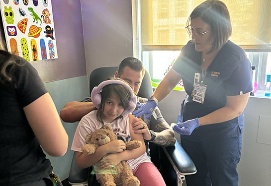 A young girl wearing fuzzy pink headphones sits in a medical chair on her father's lap while two health care providers prep her for a vaccine.