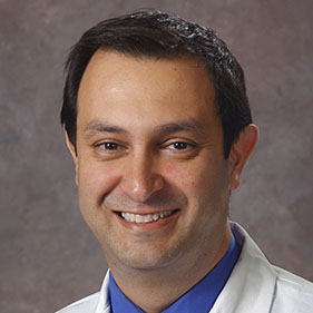 Headshot of a dark haired man in with a blue collar and a white doctor's coat.