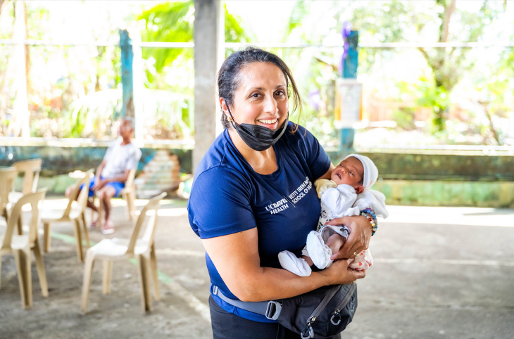 Nurse with a face mask under her chin smiles while holding a newborn baby in a community clinic setting.