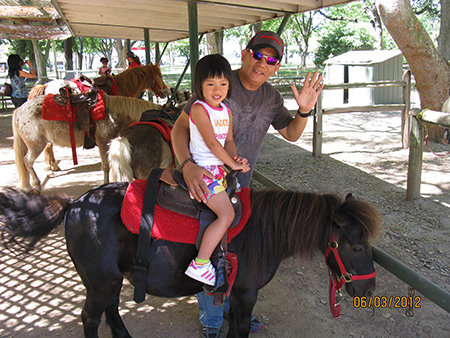 Asian man and young girl in a horse stable where she sits on a saddle while he waves at the camera wearing sunglasses.
