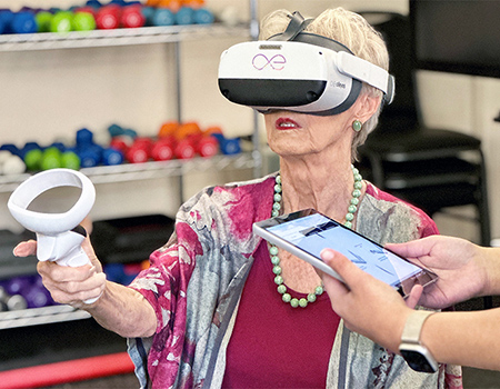 A senior woman wearing a virtual-reality headset raises a controller in her right hand.