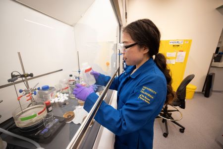 a women with safety goggles and a blue lab coat works in a lab with chemicals