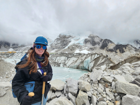 A hiker stands on a rocky trail with trekking poles, overlooking a dramatic glacier and snow‑covered mountains under heavy clouds.