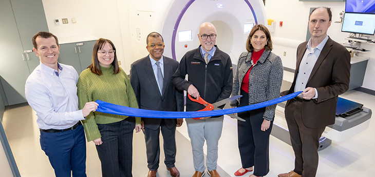 Five people in a room stand next to man with large scissors at ribbon-cutting ceremony with a cylindrical machine nearby.