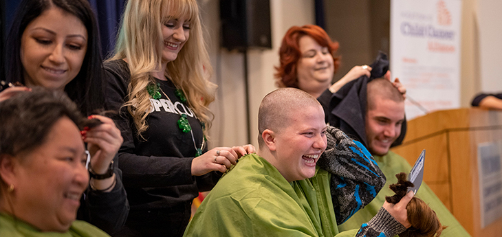 Three people on stage wearing green drapes have their heads shaved bald by three women and emcee in distance.
