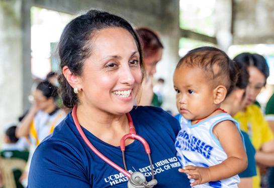 Nurse wearing a stethoscope smiles while holding a young child in a community health setting.