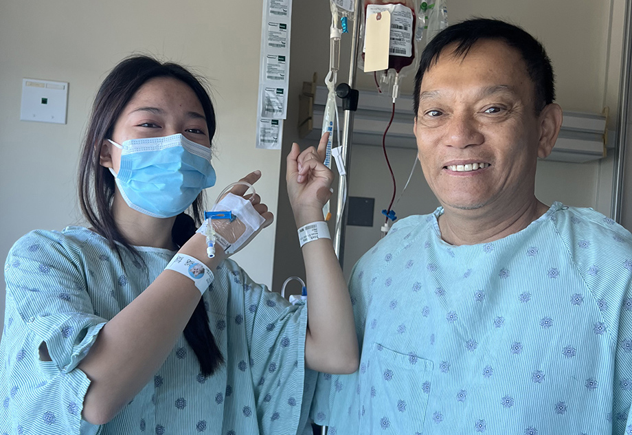 Daughter with a mask on and hospital gown stands next to her father with a hospital gown in his room after their individual bone marrow procedures..