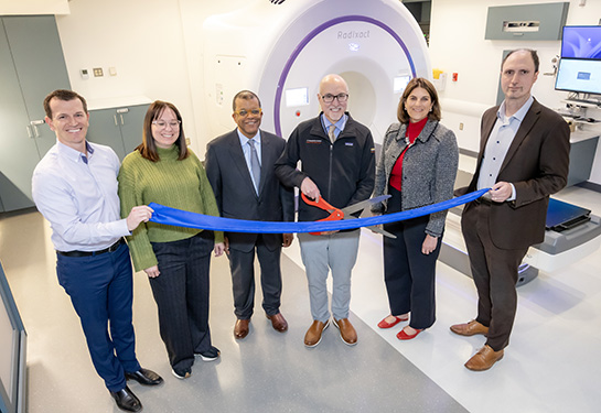 Five people in a room stand next to man with large scissors at ribbon-cutting ceremony with a cylindrical machine nearby.