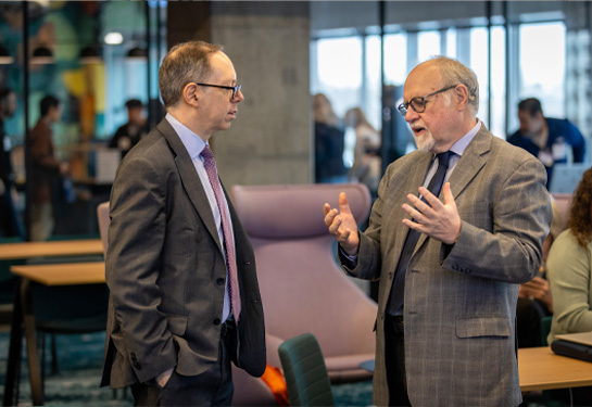 Two men in suits stand in a modern indoor space, engaged in conversation, with one gesturing as others work in the background