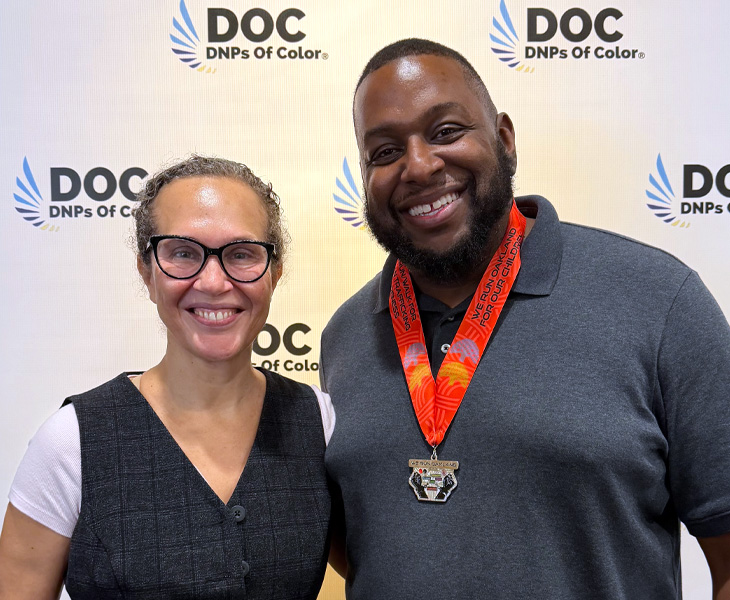 Two people smile in front of a DNPs of Color backdrop, one wearing a medal.