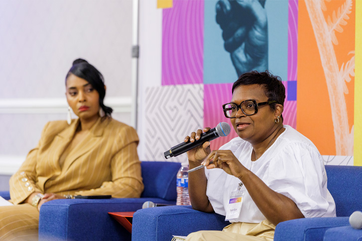 Woman speaking into a microphone during a panel discussion, with another woman seated beside her on stage.