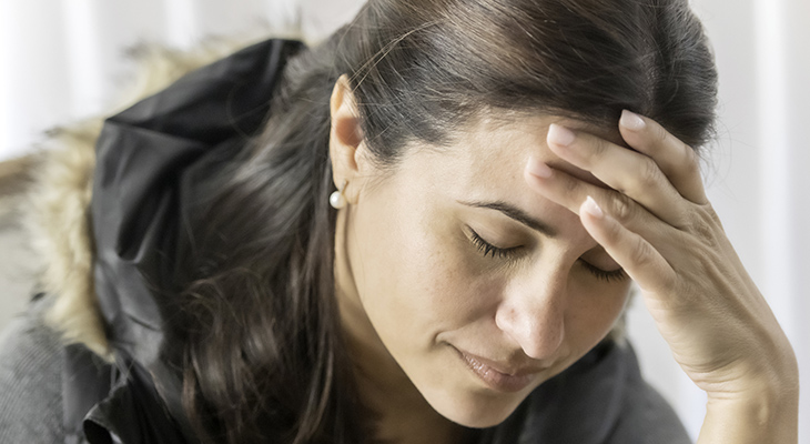 : A woman placing her hand on her forehead with her eyes closed. She is wearing pearl earrings.