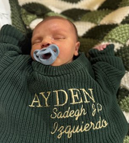 Sleeping baby boy wearing a green sweater with his name Ayden Sadegh JP Izquierdo on it.