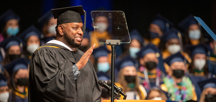 Graduate in cap and gown speaks at a podium, raising his hand beside a teleprompter, with masked graduates seated behind him.