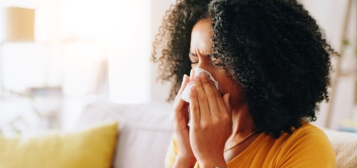 Person on a couch blowing nose with a tissue in a bright living room.