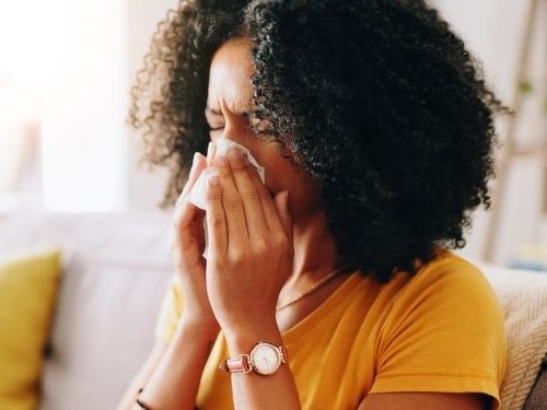 Person on a couch blowing nose with a tissue in a bright living room.