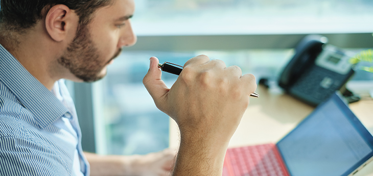A man in a light blue shirt holding a pen working on his laptop