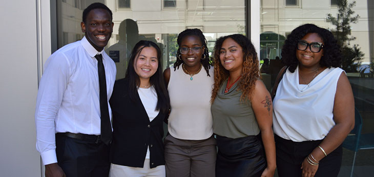 A group of five college students, all MINDGAP scholars, stand outside for a group photo next to a building lined with windows.