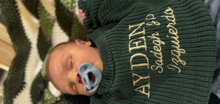 Sleeping baby boy wearing a green sweater with his name Ayden Sadegh JP Izquierdo on it.