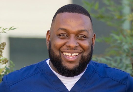 Nurse in navy blue medical scrubs smiles in outdoor area where greenery is softly blurred in the background