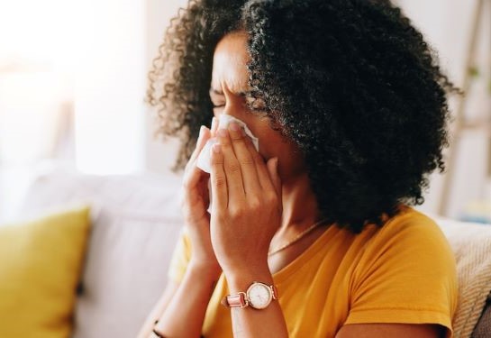 Person on a couch blowing nose with a tissue in a bright living room.