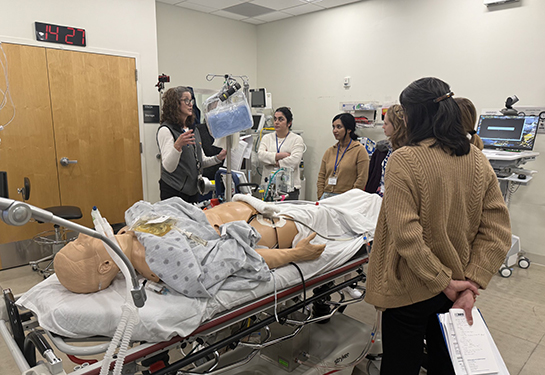 A woman gestures with hands while speaking to five people gathered around an adult sized mannequin on a gurney.