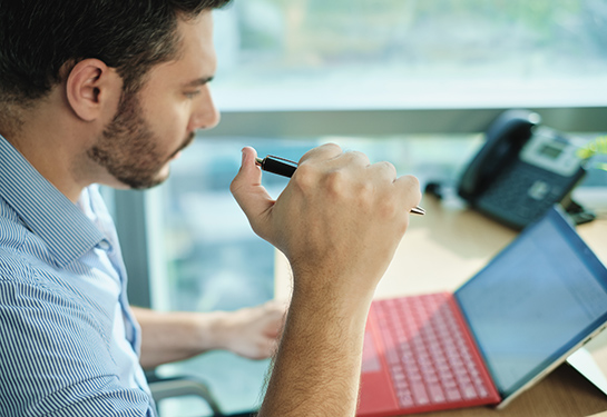 A man in a light blue shirt holding a pen working on his laptop