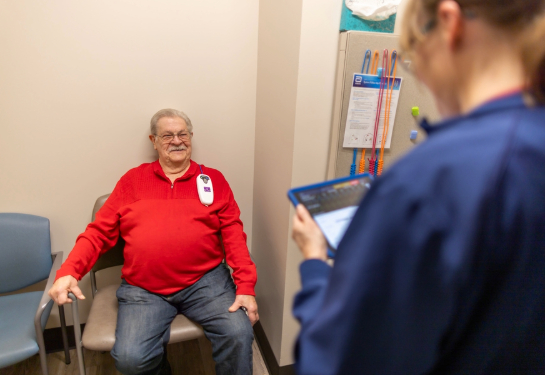 An older adult sits in a medical exam room chair while a healthcare worker stands nearby reviewing information on a tablet, with clinical equipment an