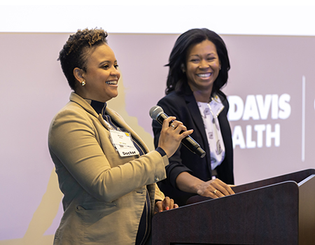 Two women at a podium with large screen behind them that reads 