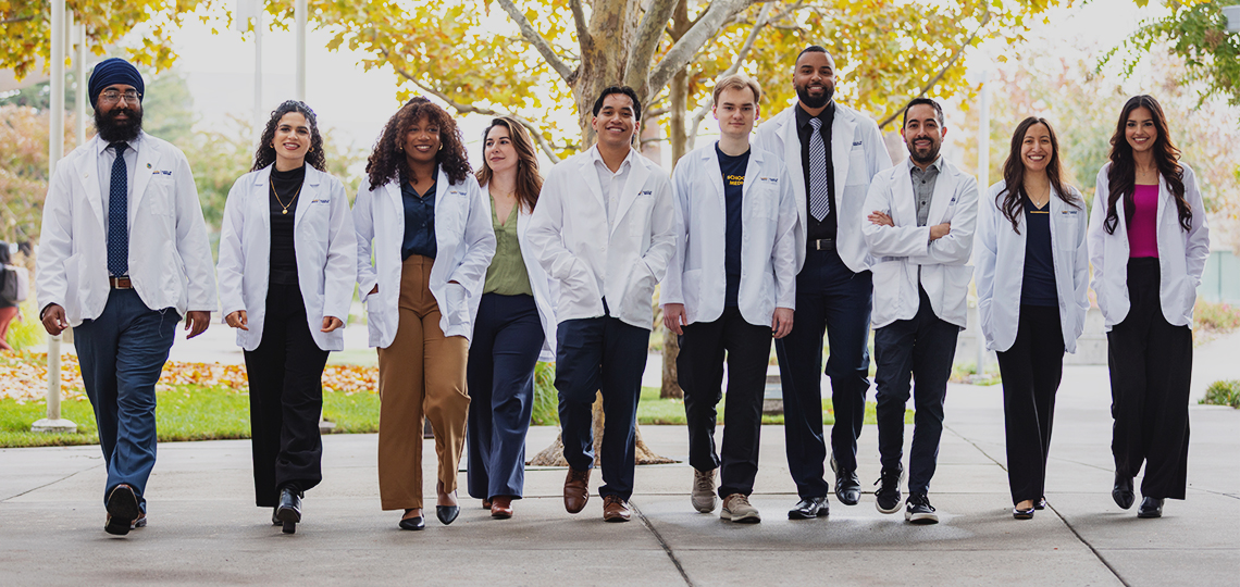 A group of 10 students, most in white coats, stand side by side as they walk in a courtyard at the UC Davis School of Medicine.